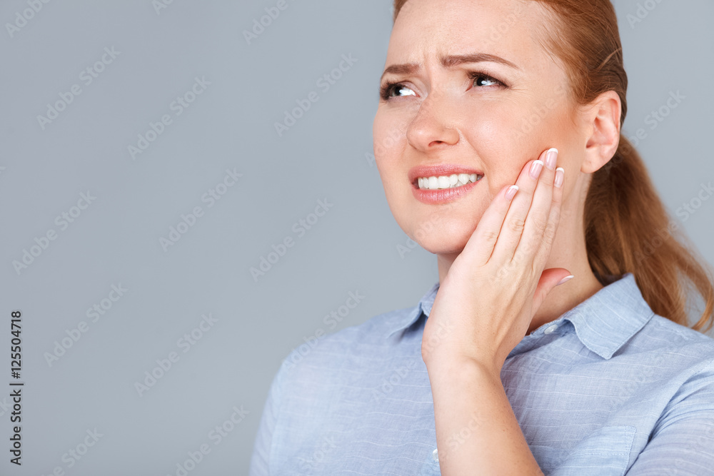 Suffering from toothache. Beautiful young woman touching her cheek standing against grey background