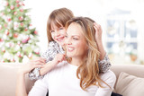 Mother and daughter at christmas. Close-up portrait of cute little girl hugging her happy mother while sitting at home in christmas time. 