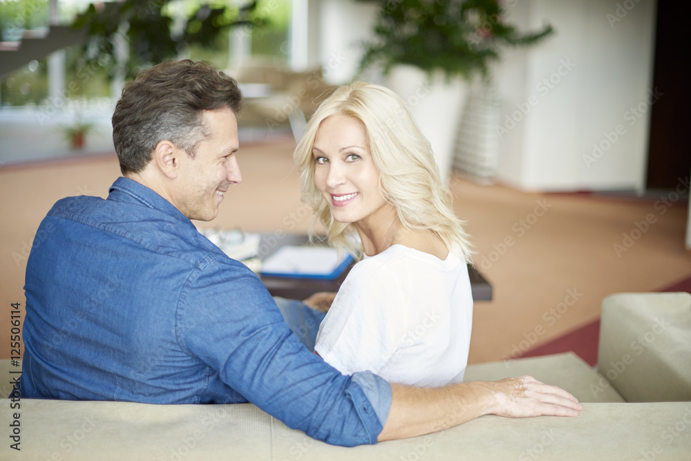 Quality time together. Rear view shot of a beautiful middle aged couple sitting on sofa at home and relaxing. Beautiful woman looking back while her husband hugging her. Quality time together. Rear view shot of a beautiful middle aged couple sitting on sofa at home and relaxing. Beautiful woman looking back while her husband hugging her.