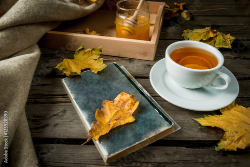 Cozy autumnal mood, warm autumn. A cup of hot tea with lemon and ginger on a rustic table, plaid, yellow leaves, honey and a book to read.