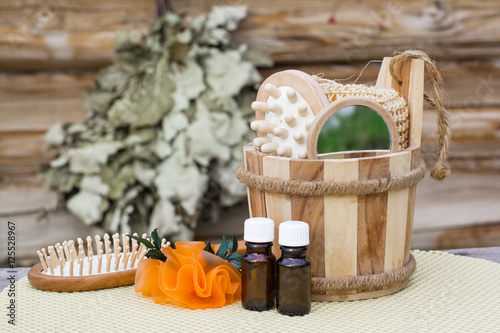Деревянный ковш с предметами для купания, эфирные масла, мыло, расческа на фоне банного веника photo