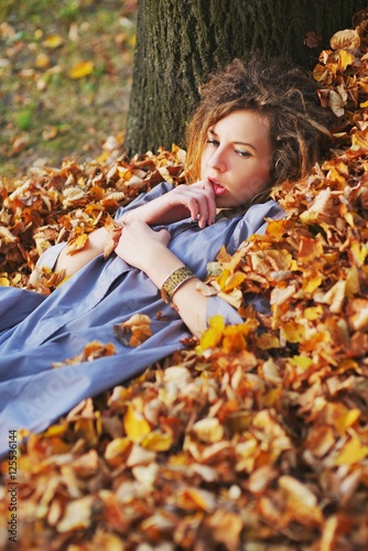 Cute teen girl in a light purple cloak with sad eyes lies on a pile of fallen autumn leaves in the forest  leaning his head against the tree  close up.