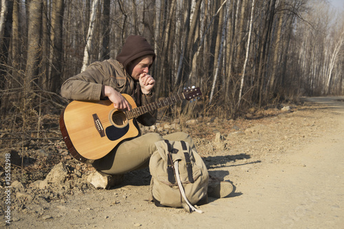 The hooded man with a backpack and guitar sitting beside the road and your hands warm breath