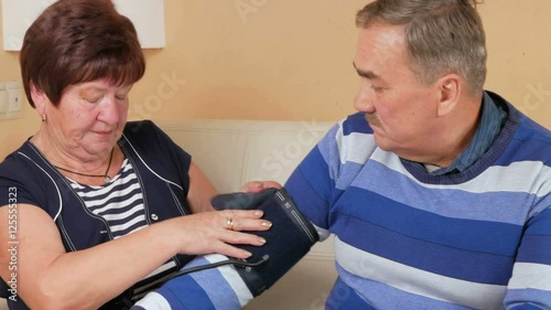 Elderly woman measures the blood pressure at home on the couch. Poor man's well-being. Taking care of his wife about her husband photo