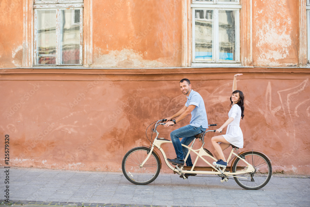 Naklejka premium Happy couple riding on retro tandem bicycle at the street city against the background of the old orange wall with windows. The man runs a bicycle, a girl in white dress raised her hand up. Lviv