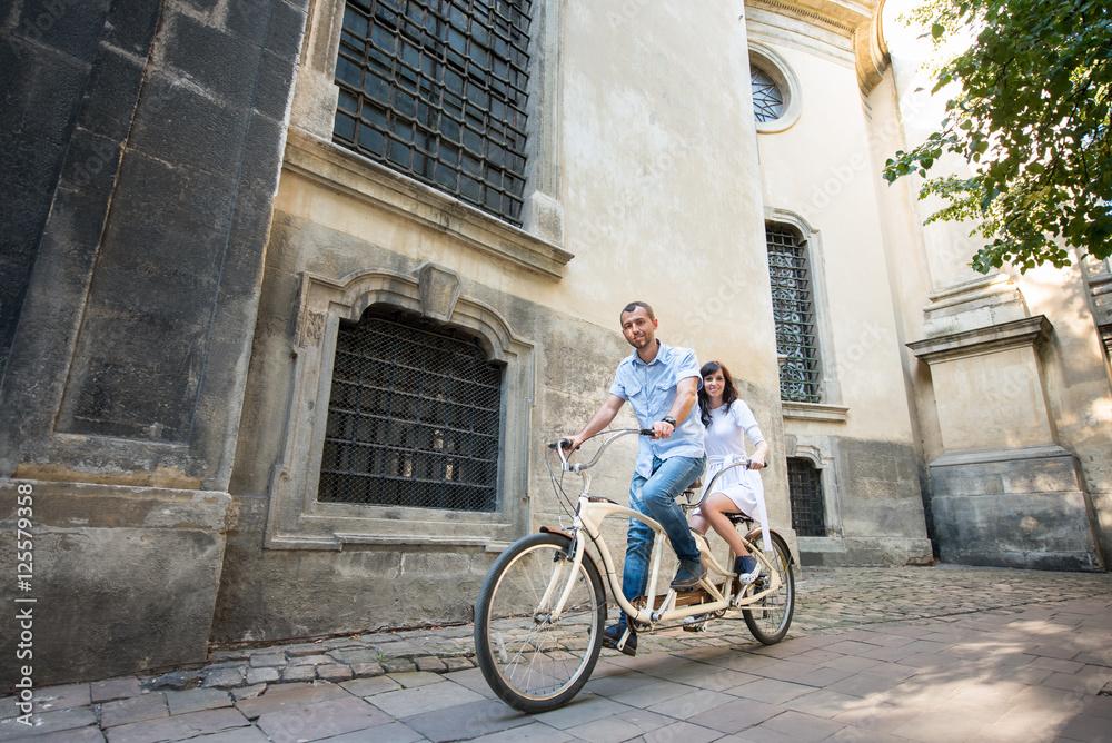 Happy couple riding on retro tandem bike against the background of historic building at the sunny day. Guy in jeans and a shirt and a girl in a white dress. Lviv, Ukraine