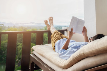Rear view of asian man relaxing on a sofa and holding book on bed at home terrace with beautiful green background view. Relaxing concept.