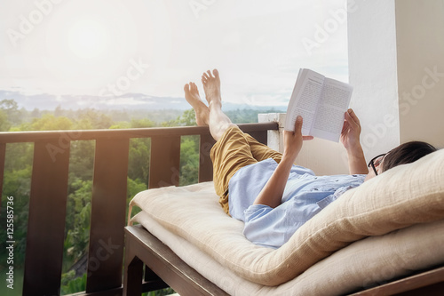 Rear view of asian man relaxing on a sofa and holding book on bed at home terrace with beautiful green background view. Relaxing concept.