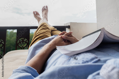 Young man reading a book lying on soft mattress in relaxing bed at terrace with green nature view. Fresh air in the morning of weekend or free day. Relax or education background idea. Selective focus. photo