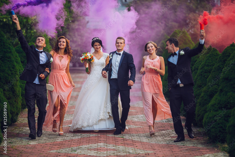 Newlyweds and their friends raise their hands up posing on the lawn. Running with smoke bombs.
