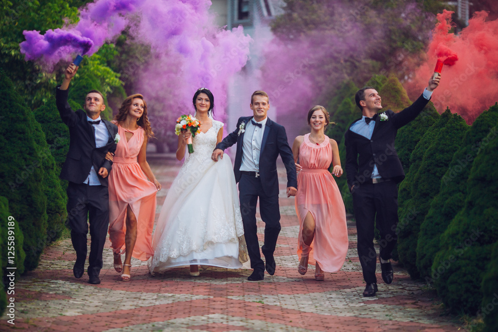 Newlyweds and their friends raise their hands up posing on the lawn. Running with smoke bombs.