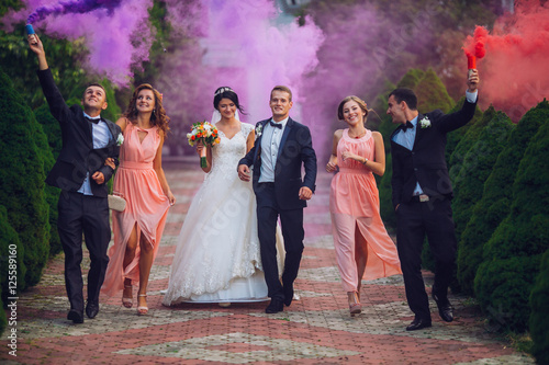 Newlyweds and their friends raise their hands up posing on the lawn. Running with smoke bombs.