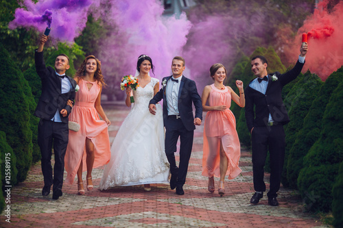 Newlyweds and their friends raise their hands up posing on the lawn. Running with smoke bombs.