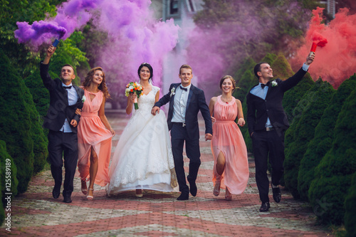 Newlyweds and their friends raise their hands up posing on the lawn. Running with smoke bombs.