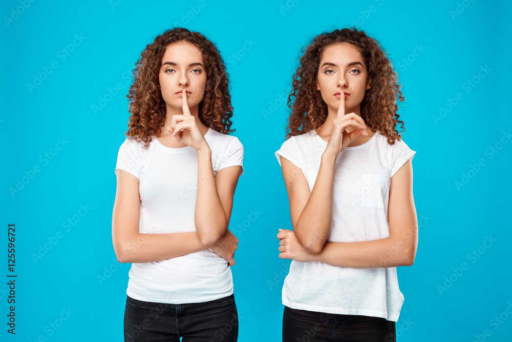 Two pretty girls twins showing keep silence over blue background.