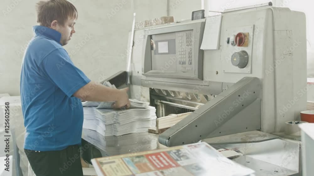 Worker in blue jacket worker puts the paper in the cutter guillotine machine in a printing factory, slider Worker in blue jacket worker puts the paper in the cutter guillotine machine in a printing factory, slider