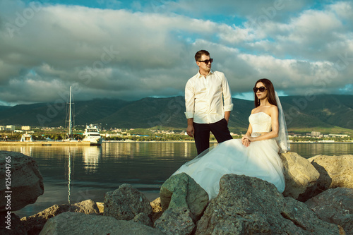 Portrait of young just married couple in wedding gowns and stylish sunglasses on the rock at the seaside.  Mountains on background. Wedding fashion and eyewear concept. Copy-space. Outdoor shot © obrik