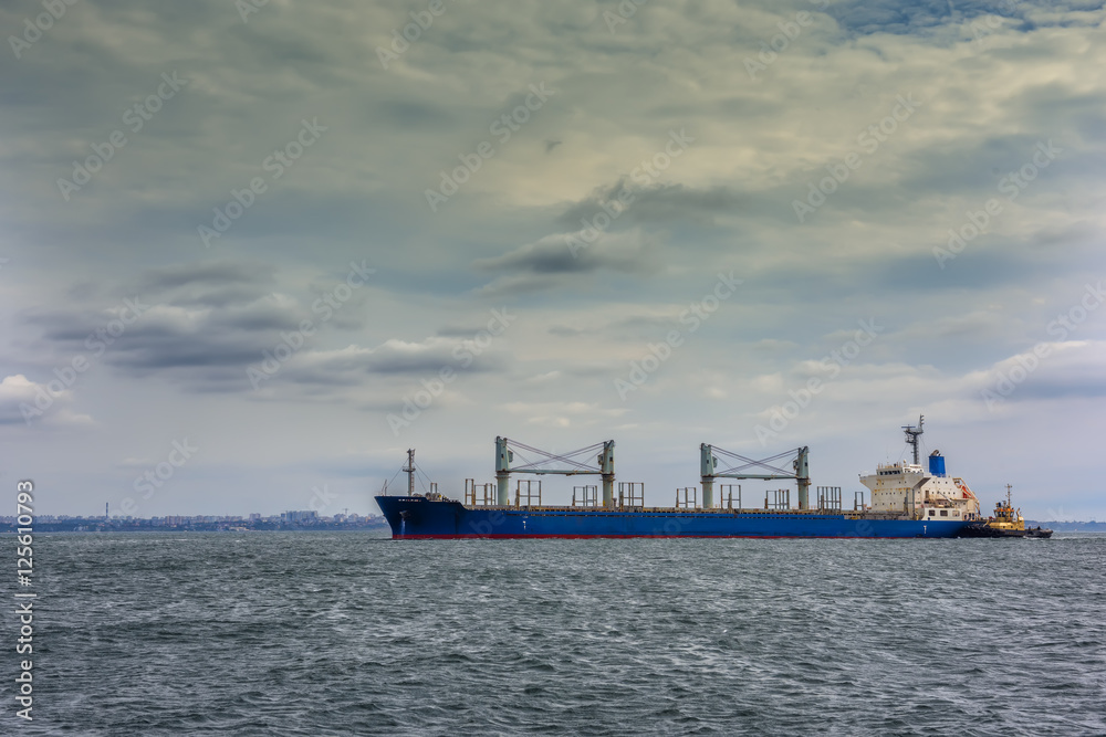 large ship cargo ship with cranes in the sea followed by a tug to the shore. In the background visible mnogoetyzhnye city building. Beautiful sky with clouds. Overcast
