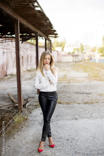 Beautiful tall girl with long blonde hair and red lipstick. Dressed in a white fashionable shirt tucked in black latex pants, on her legs are red stilettos. Modern office woman