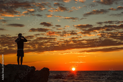 Hipster guy taking pictures of amazing landscape on mobile smart phone digital camera while standing on a rock near the sea, beautiful sunset light create background with copy space for text message