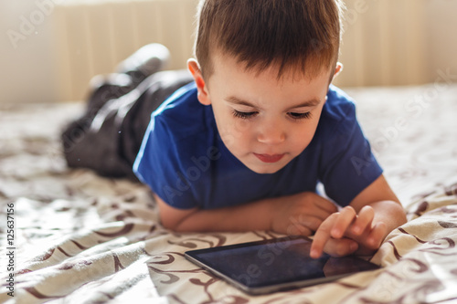 Little boy using tablet while lying on bed, natural light.