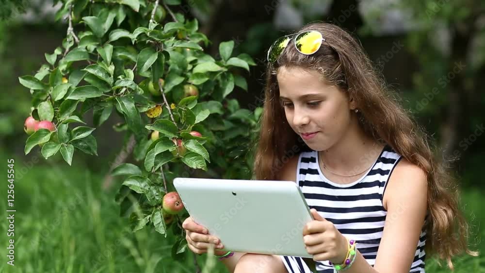 Pretty girl in sunglasses with tablet pc stands under apple tree in garden. Female in glasses with tablet pc. Girl communicates through skype. Nice girl stands under apple tree and talks via tablet pc