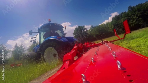 A blue tractor is driving across a big meadow and it is cutting crass with a grass cutting machine. Close-up shot. It is a nice summer day.
