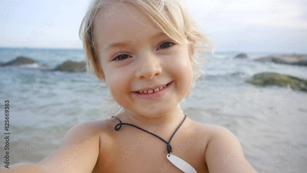 A cute little child video chats on smart phone on the beach on blue sea background. Using  a portable handheld device on summer vacation. Talking on a mobile. Doing selfie. Wide angle view