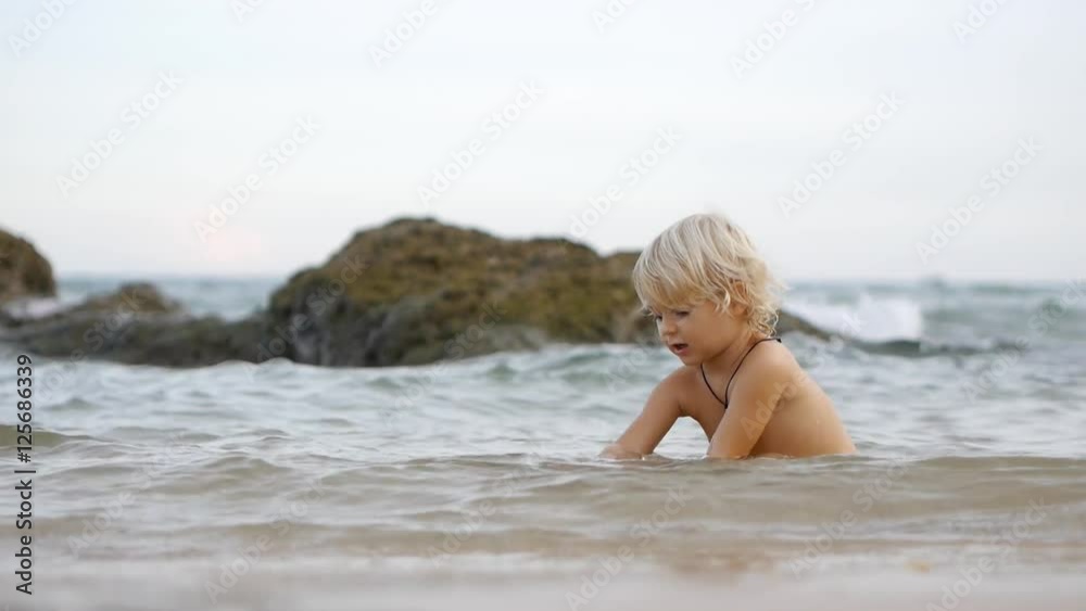 Little cute smiling child playing with shells and stones on waves in blue sea, waving hand. Having fun in summer vacation. Static view Little cute smiling child playing with shells and stones on waves in blue sea, waving hand. Having fun in summer vacation. Static view