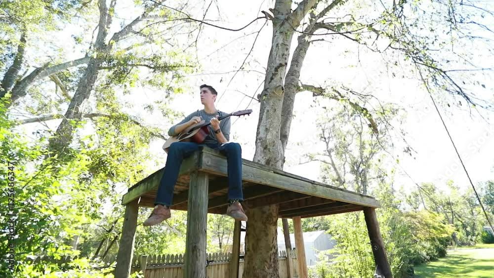 Teenage boy plays guitar on a tree platform, slow motion backlit by bright afternoon sun. Teenage boy plays guitar on a tree platform, slow motion backlit by bright afternoon sun.