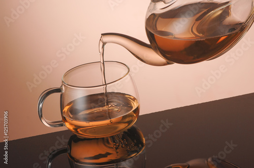 Transparent glass teapot and cup of tea on the reflective surface