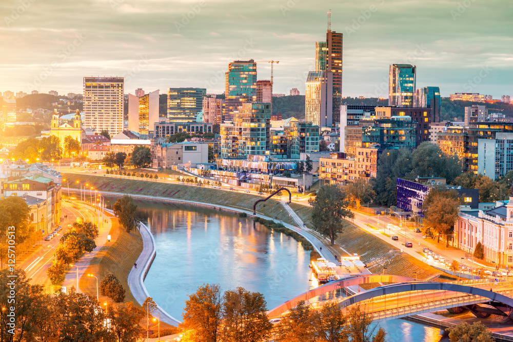 Fototapeta premium Cityscape view on the illuminated financial district with modern buildings, river and bridge during the sunset in Vilnius, Lithuania.
