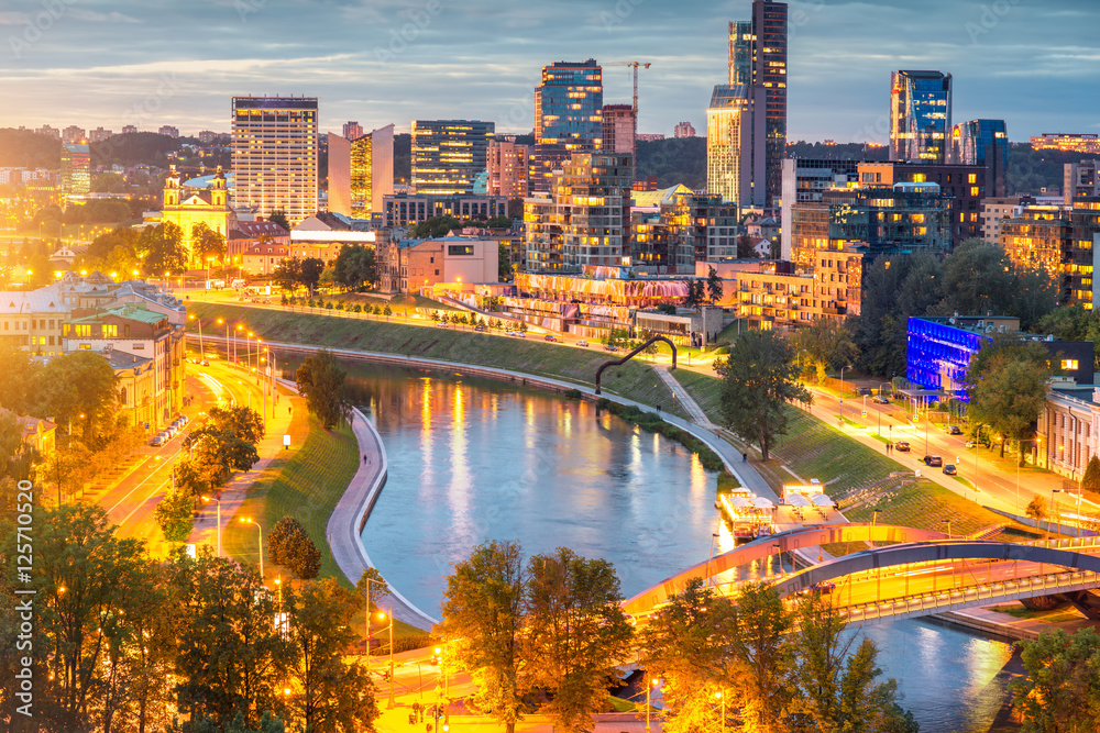 Fototapeta premium Cityscape view on the illuminated financial district with modern buildings, river and bridge during the sunset in Vilnius, Lithuania.