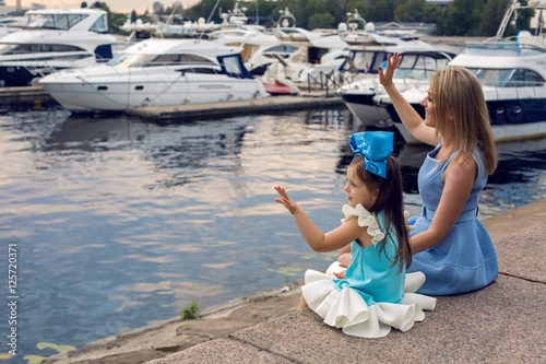 little girl three years sitting with mom with long blond hair and they are dressed in blue dress and large blue bow on her head , sitting at the pier on the sea with a yacht during sunset © saulich84