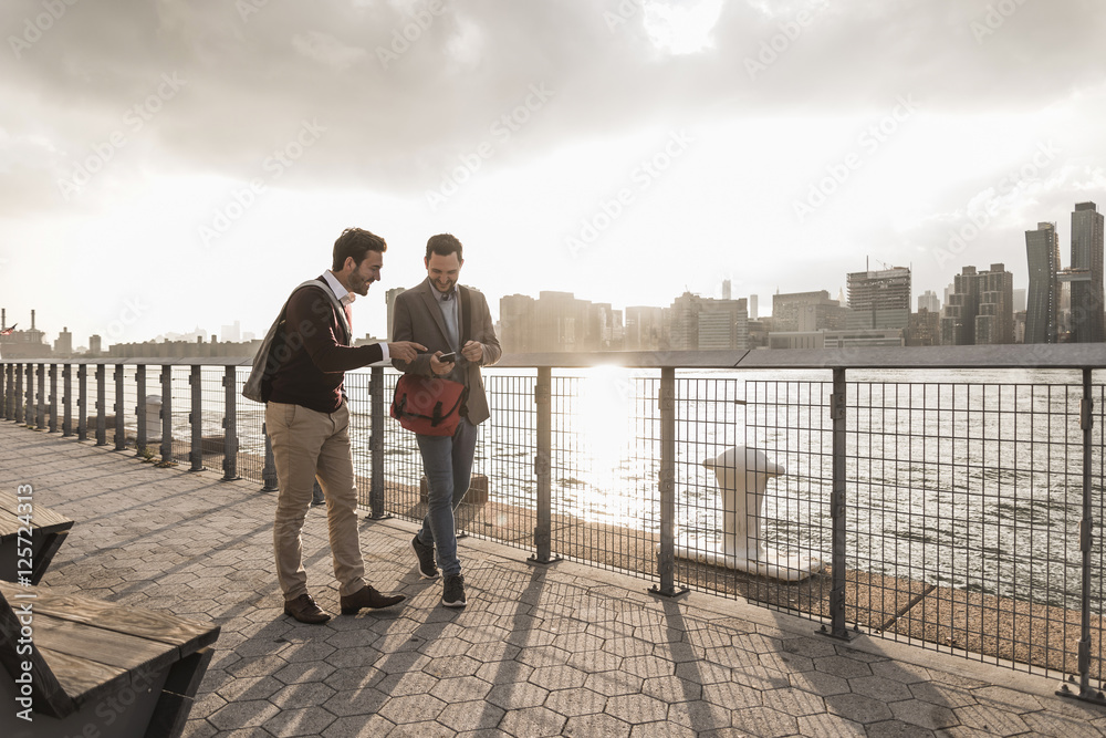 USA, New York City, two young men walking along East River looking at cell phone USA, New York City, two young men walking along East River looking at cell phone