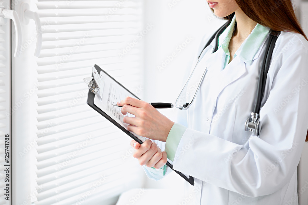 Female doctor filling up medical history standing near the window in hospital Female doctor filling up medical history standing near the window in hospital