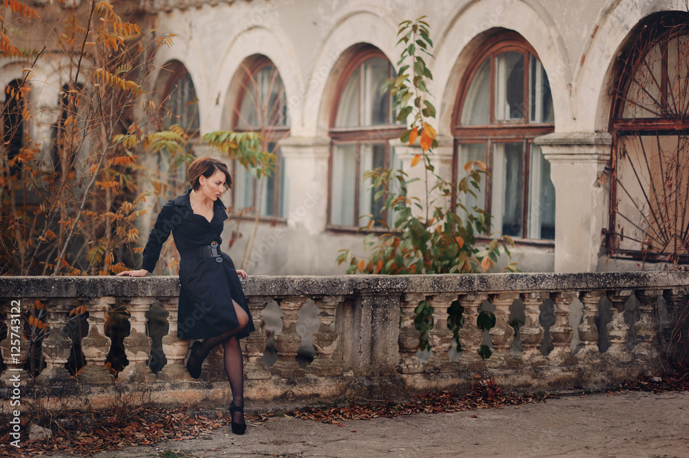 Молодая женщина в черном пальто стоит возле заброшенного старинного здания с колоннами 