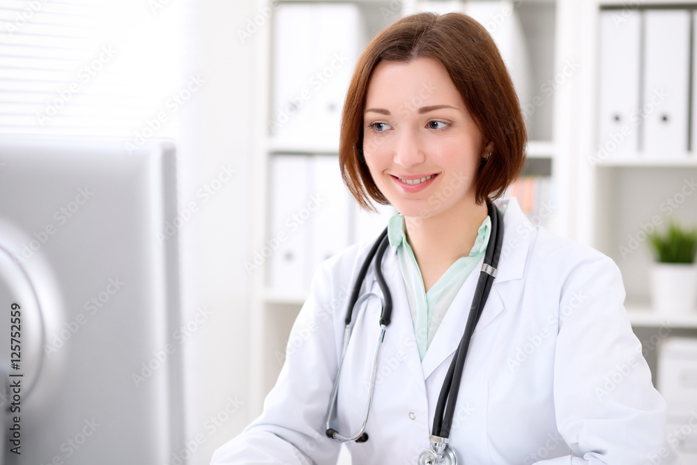 Young brunette female doctor sitting at a desk and working on the computer at the hospital office. Health care, insurance and help concept. Physician ready to examine patient Young brunette female doctor sitting at a desk and working on the computer at the hospital office. Health care, insurance and help concept. Physician ready to examine patient