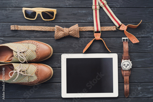 Outfit of male or female traveler, student, teenager, hipster. Top view of essentials for modern young person. Tablet, sunglasses, shoes, watch, bow tie and suspenders on black wooden background