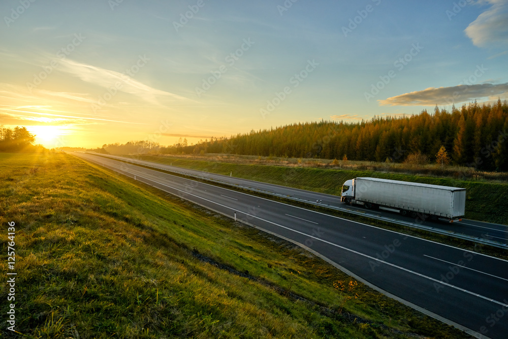 Asphalt highway with a ride white truck between green meadows and larch forest in autumn landscape at sunset.