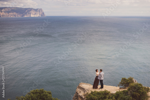 Romantic and stylish caucasian couple standing on the background of spectacular sea view. Love, relationships, romance, happiness concept.