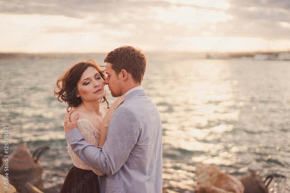 Romantic and stylish caucasian couple hugging at sunrise on the rocks near the sea. Love, relationships, romance, happiness concept.
