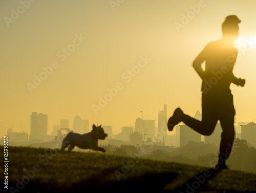 Defocus silhouettes of jogger and his dog run in front of the golden sunrise city skyline in London