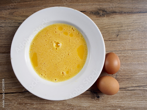 Beaten egg in old plate with two eggs on wood background