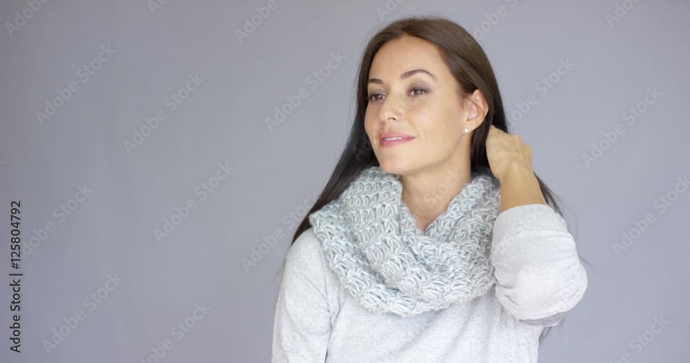 Elegant beautiful middle aged woman posing with woolen warm scarf around her neck. She smiling and trying to warm up herself. Isolated on gray background.