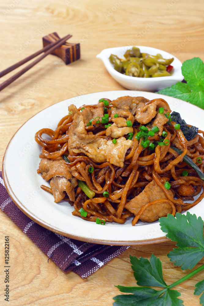 yi mein noodle stir fried with dark caramel sauce on wooden table, served together with a small plate of sour taste green chili, makes a delectable authentic malaysian chinese street food. yi mein noodle stir fried with dark caramel sauce on wooden table, served together with a small plate of sour taste green chili, makes a delectable authentic malaysian chinese street food.