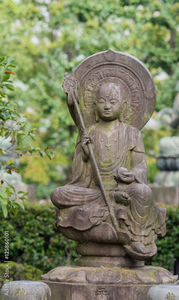 Tokyo, Japan - September 26, 2016: Closeup of stone statue of Bodhisattva sitting on lotus pedestal in garden at Senso-ji Buddhist Temple. Large halo. Holds staff and sphere. Green foliage. 