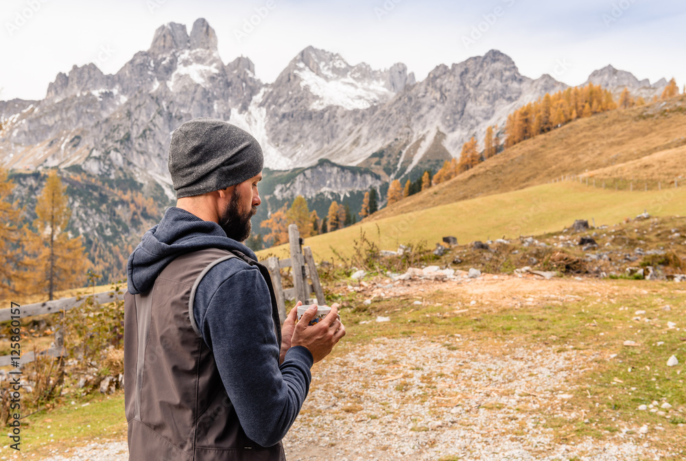 Bearded hiking man in Alps mountains drinking tea or coffee and enjoying amazing view of autumn nature. Traveler warming his hands with mug of tea. Alps Austria Europe at fall season. 