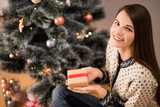 Young beautiful woman in knitted sweater holding gift in hands and sitting near decorated Christmas tree, New Year concept