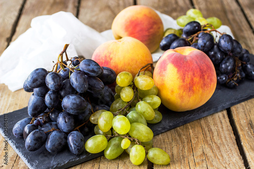 A bunch of grapes and ripe peaches on a black slate board on a wooden table